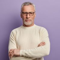 Headshot of confident mature European male pensioner keeps arms folded, wears optical glasses and white sweater, being sure in something, isolated over purple background. People and aging concept